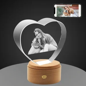 A heart-shaped crystal with a 3D laser-engraved photo of a long-haired woman hugging her Labrador, set on a round wooden rotating LED base, with the original color photo displayed on a smartphone above, against a gray background.