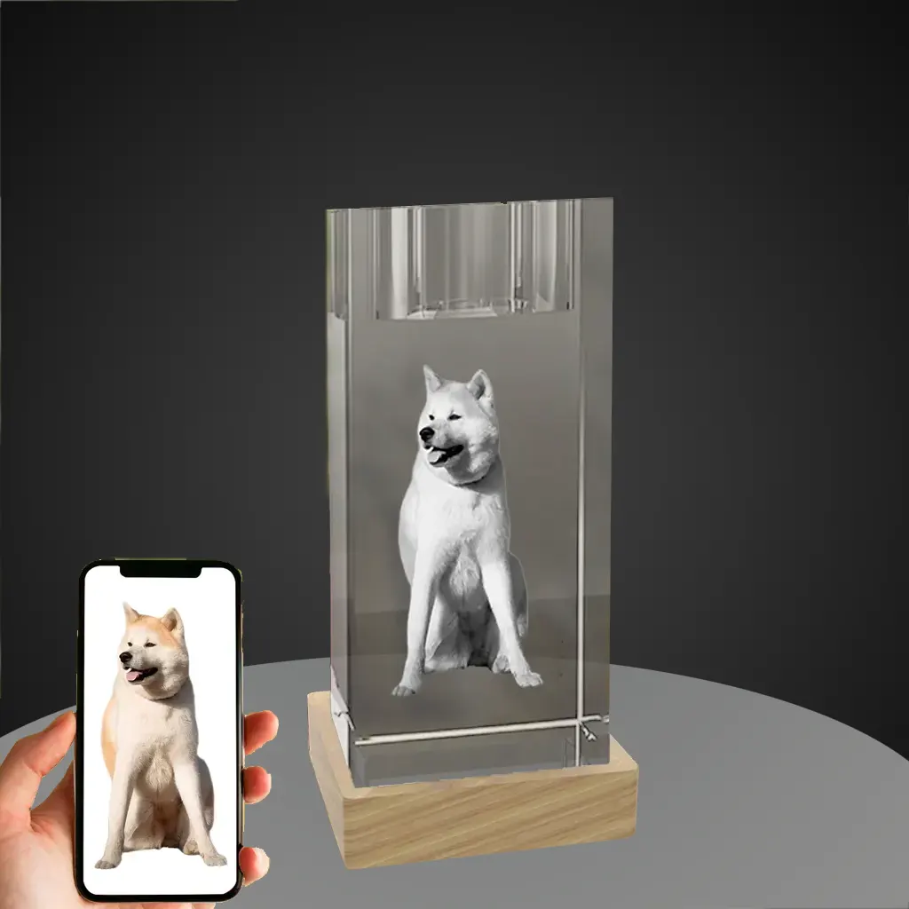 A hand holds a smartphone showing a color photo of an Akita dog, next to a 3D laser-engraved candlestick crystal featuring the same dog in monochrome, resting on a wooden LED base against a gray background.