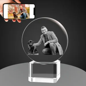 A hand holds a smartphone showing a color photo of a smiling man with his black pug, below is a crystal ball with a 3D monochrome engraving of the same scene, resting on a clear, square crystal base against a gray background.