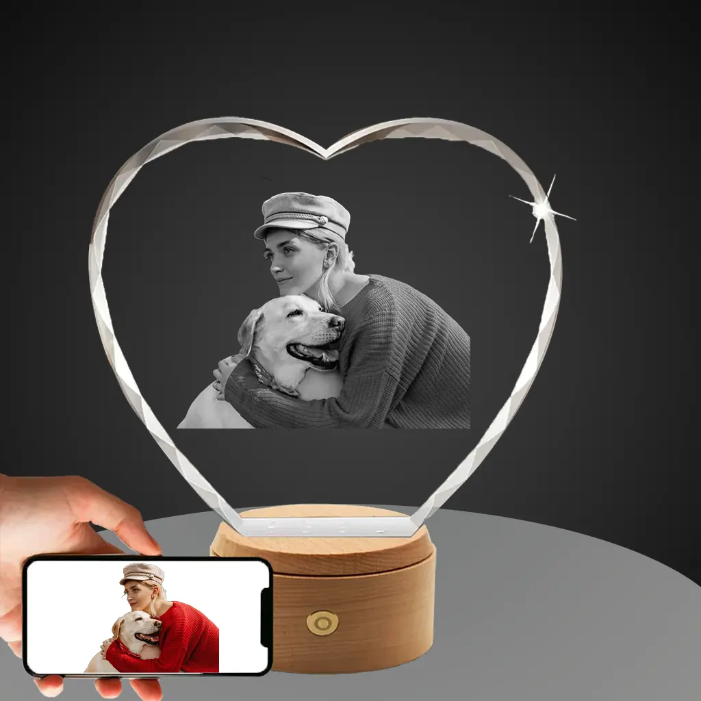 A hand holds a smartphone showing a color photo of a woman hugging her Labrador, next to a diamond-cut heart-shaped crystal with the same image 3D-engraved in monochrome, resting on a round, wooden, rotating LED base.