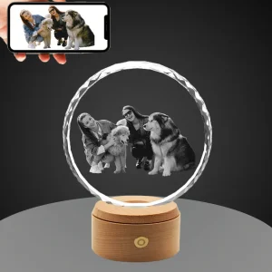 A hand holds a smartphone showing a color photo of two women and two pet dogs, below which is a sunflower-shaped, round, diamond-cut crystal with the same photo 3D-engraved in monochrome, placed on a round wooden LED base against a gray background.