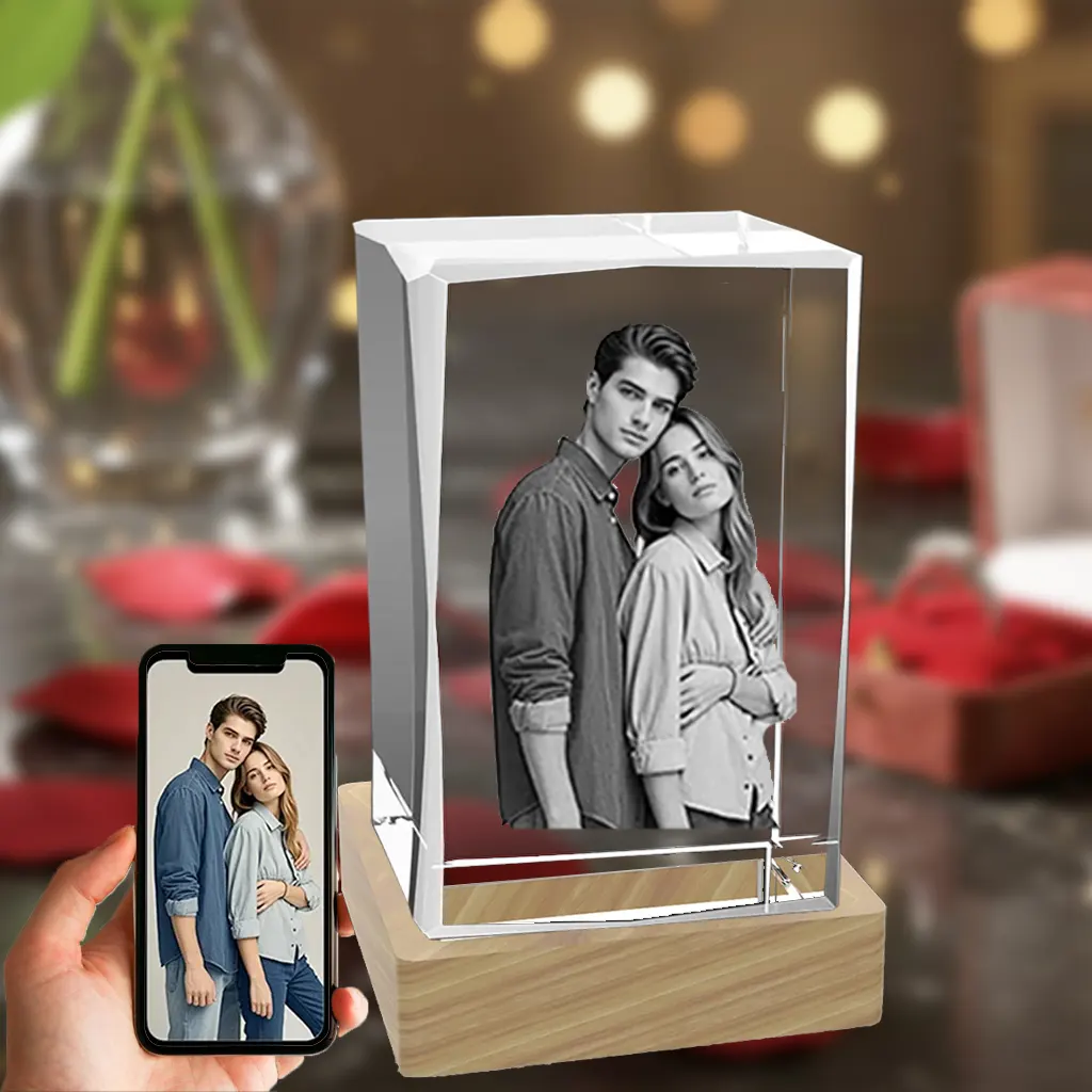 A beveled rectangular 3D crystal internally engraved with an intimate portrait of a couple. The crystal sits on an illuminated wooden LED base, against a romantic background of red roses and gifts. In the bottom-left corner, a hand holds a smartphone displaying the original color photograph.