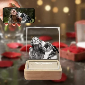 A beveled rectangular 3D crystal internally engraved with a heartwarming picture of a woman and her pet dog sharing a kiss, with the text "Our Soulmate" below. The crystal sits on an illuminated wooden LED base, against a romantic red rose background, with a hand in the top-left holding a phone showing the original color photo.