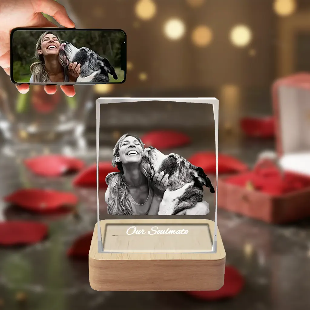 A beveled rectangular 3D crystal internally engraved with a heartwarming picture of a woman and her pet dog sharing a kiss, with the text "Our Soulmate" below. The crystal sits on an illuminated wooden LED base, against a romantic red rose background, with a hand in the top-left holding a phone showing the original color photo.