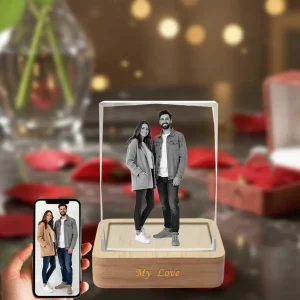 A vertical, beveled rectangular 3D crystal internally engraved with a full-body portrait of a couple, placed on an illuminated wooden LED base engraved with "My Love." The background is a romantic scene with red roses, and a phone in the bottom-left corner displays the original color photograph.