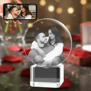 A crystal ball with a 3D engraved portrait of a couple looking lovingly at each other, resting on a square crystal base. The background is a romantic scene with red roses and gifts, and a hand in the top-left corner holds a phone displaying the original color photo.