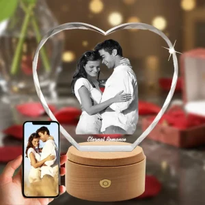A brilliant, diamond-cut heart-shaped 3D crystal, internally engraved with a romantic portrait of a hugging couple and the text "Eternal Romance". The crystal is set on a rotating, illuminated wooden LED base against a romantic red rose background, with a phone in the bottom-left showing the original color photo.