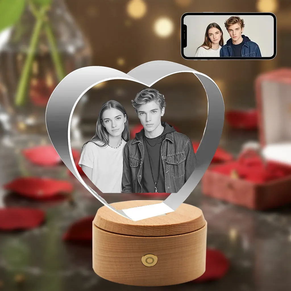 A heart-shaped 3D crystal internally engraved with a portrait of a young couple, placed on a rotating, illuminated round wooden LED base. The background is a romantic scene with red roses and a gift box, and a smartphone in the top-right corner displays the original color photo.