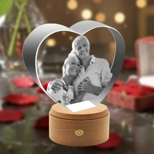 A heart-shaped 3D crystal internally engraved with a loving portrait of an elderly couple, placed on a rotating, illuminated round wooden LED base against a romantic background of red roses and gift boxes.