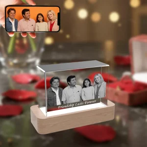 A horizontal rectangular 3D crystal engraved with a four-person group photo and the text "Friendship Lasts Forever," placed on an illuminated wooden LED base. The background is a romantic scene with red roses and gifts, and a hand in the corner holds a phone showing the original color photo.