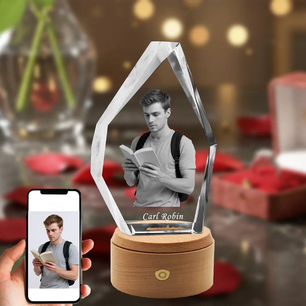 An iceberg-shaped 3D crystal engraved with a young man reading a book with a backpack on, and the name "Carl Robin" etched below. The crystal is set on a rotating, illuminated wooden LED base against a romantic red rose background, with a hand in the bottom-left holding a phone showing the original photo.