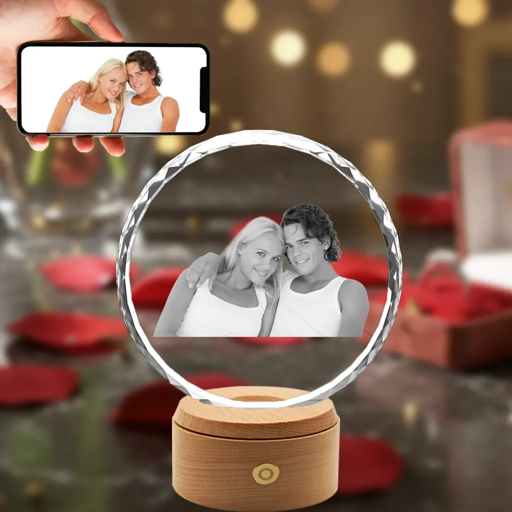 A sunflower-shaped 3D crystal internally engraved with an intimate portrait of a young couple. The crystal sits on an illuminated round wooden LED base against a romantic background of red roses, with a hand in the top-left holding a smartphone displaying the original color photo.
