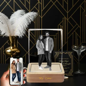 A 3D photo of a couple is engraved in a rectangular crystal, which sits on a wooden LED base engraved with "My Love". A hand holds a phone showing the original color photo, against an art deco background with a vase of feathers and a champagne glass.