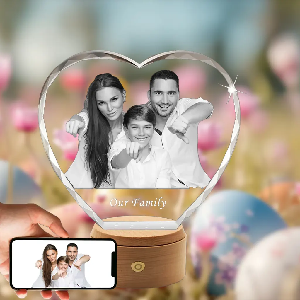 A 3D photo of a smiling family is engraved in a faceted, heart-shaped crystal with 'Our Family' text. The crystal rests on a wooden rotating LED base, set against a blurry background of Easter eggs and flowers, with a hand holding a phone displaying the original photo.