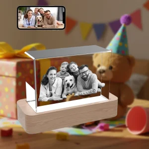 A personalized horizontal rectangular 3D photo crystal gift. Inside is a laser-etched photo of a happy family with their dog. The crystal rests on a wooden LED base. A smartphone above shows the original color photo, set against a festive birthday party background.