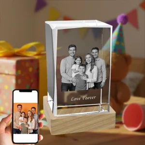 A rectangular 3D photo crystal gift on a wooden LED base, featuring a laser-etched family portrait inside. The crystal is engraved with "Love Foever." A hand holds a smartphone displaying the original color photo, set against a festive birthday party background.