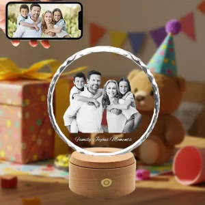 A personalized sunflower-shaped 3D photo crystal with a diamond-cut edge, featuring an engraved family photo and the text 'Family Joyous Moments'. It sits on a wooden LED base. A smartphone in the foreground shows the original color photo, set against a festive birthday party background.