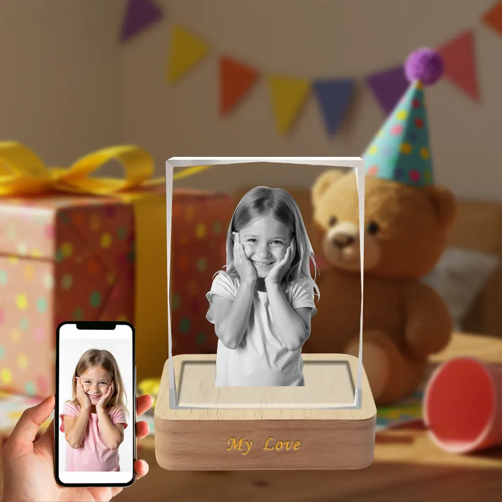 A personalized beveled-edge rectangular 3D photo crystal featuring a laser-etched image of a smiling young girl. The crystal sits on a wooden LED base engraved with 'My Love'. A hand holds a smartphone showing the original color photo, against a festive birthday party background.