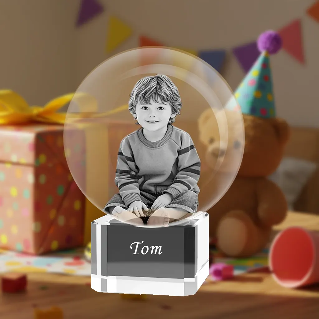 A personalized 3D photo crystal ball gift for a child. Inside the sphere is a laser-etched black and white photo of a smiling young boy. The crystal base is engraved with the name 'Tom'. The background is a blurry birthday party scene with a wrapped gift and a teddy bear in a party hat.