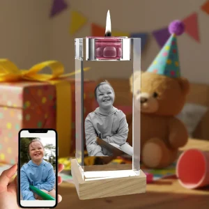A personalized 3D photo crystal candle holder with a lit candle on top. A smiling girl's photo is laser-engraved inside the crystal, which rests on a wooden base. A hand holds a smartphone showing the original color photo, set against a birthday party scene.