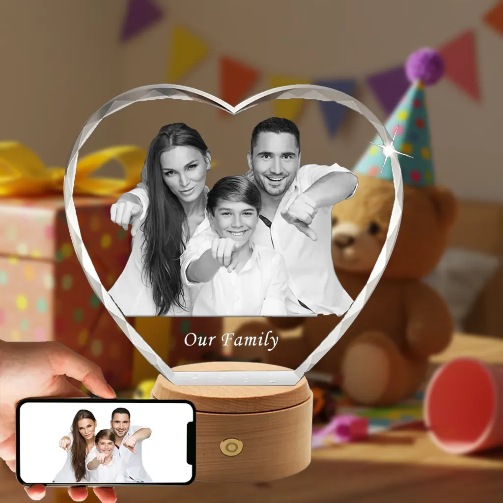 A personalized heart-shaped 3D photo crystal with a diamond-cut edge, featuring an engraved family portrait and the text "Our Family". It sits on a rotating wooden LED base. A smartphone in the foreground shows the original color photo, against a festive birthday party background.