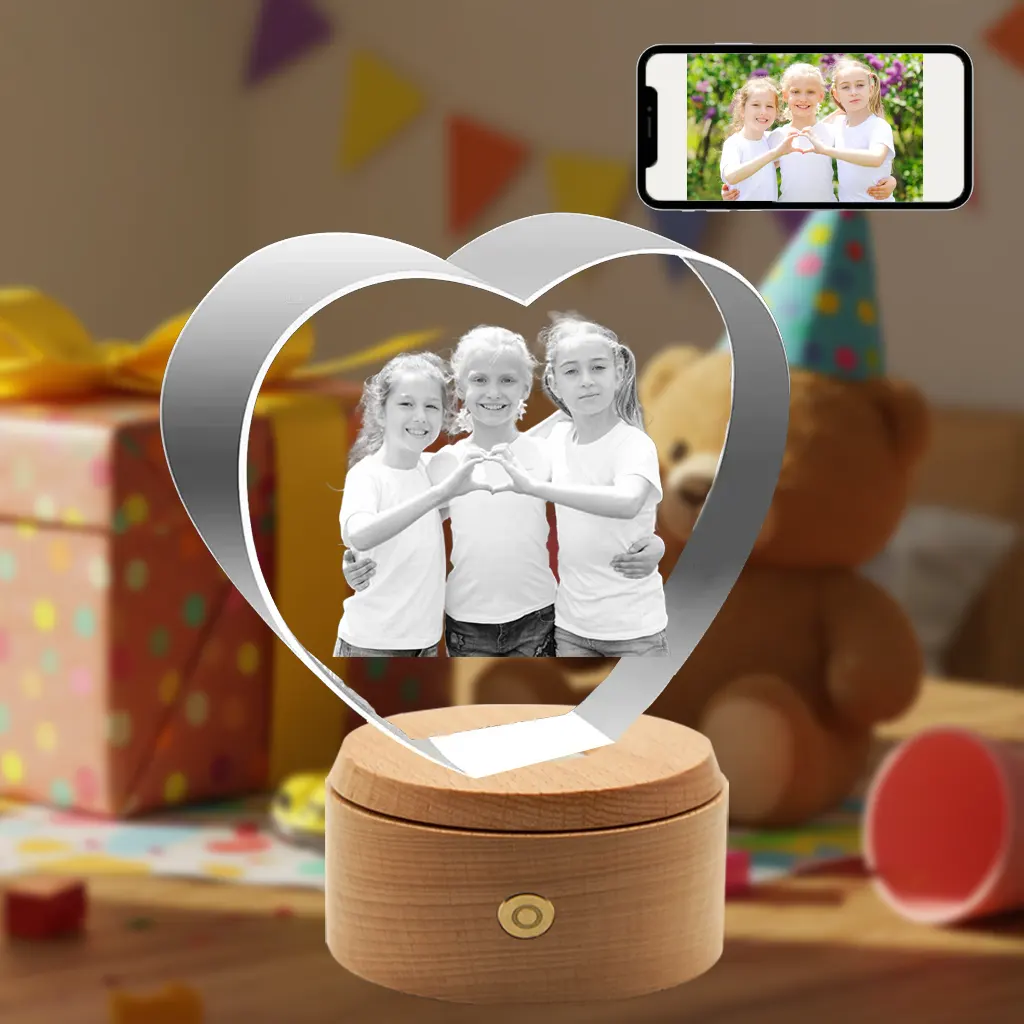 A personalized 3D photo heart-shaped crystal gift for children, featuring an engraved photo of three young girls making a heart shape with their hands. The crystal sits on a rotating wooden LED base. A smartphone displaying the original color photo is visible, set against a festive birthday party background.