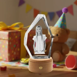A personalized iceberg-shaped 3D photo crystal gift for a child. Inside is a laser-etched photo of a smiling young girl. The crystal sits on a rotating wooden base with LED lights, against a festive background of a birthday party with a gift and teddy bear.