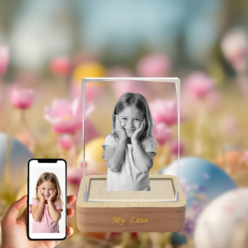 A 3D photo of a smiling little girl is engraved in a rectangular crystal, which sits on a wooden LED base engraved with "My Love". The scene is set against a vibrant Easter background of flowers and eggs, with a hand holding a phone showing the girl's original color photo.
