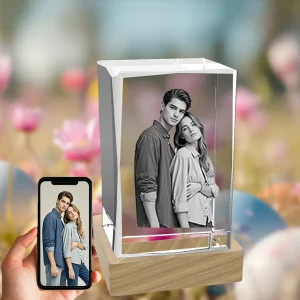A 3D photo of an embracing couple is engraved in a beveled rectangular crystal block on a wooden LED base. A hand holds a phone showing the original color photo, all set against a festive background of Easter eggs and spring flowers.