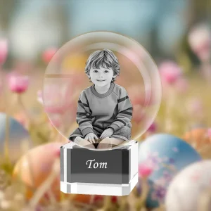 A 3D photo of a young boy is engraved inside a crystal sphere. The sphere sits on a clear crystal base with the name 'Tom' engraved on it. The background is a festive, blurry scene of Easter eggs and spring flowers.