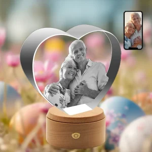 A 3D photo of a smiling elderly couple is engraved within a heart-shaped crystal, which sits on a wooden rotating LED base. The background is a festive Easter scene with flowers and eggs, and a phone in the corner shows the original color photo.
