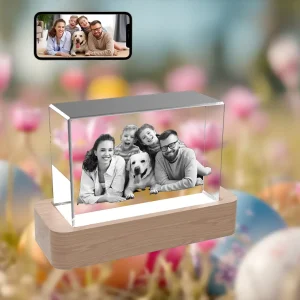 A 3D photo of a happy family with their dog is engraved in a horizontal rectangular crystal. The crystal is on a wooden LED base, against a festive Easter background of flowers and eggs. A smartphone above shows the original color family photo.