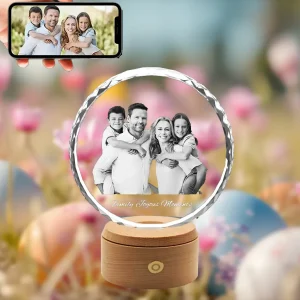 A 3D photo of a happy family, with parents giving their children piggyback rides, is engraved in a round, faceted "sunflower" crystal. The crystal, with the text "Family Joyous Moments", sits on a wooden LED base. A phone showing the original photo is held above, all against a festive Easter background of flowers and eggs.
