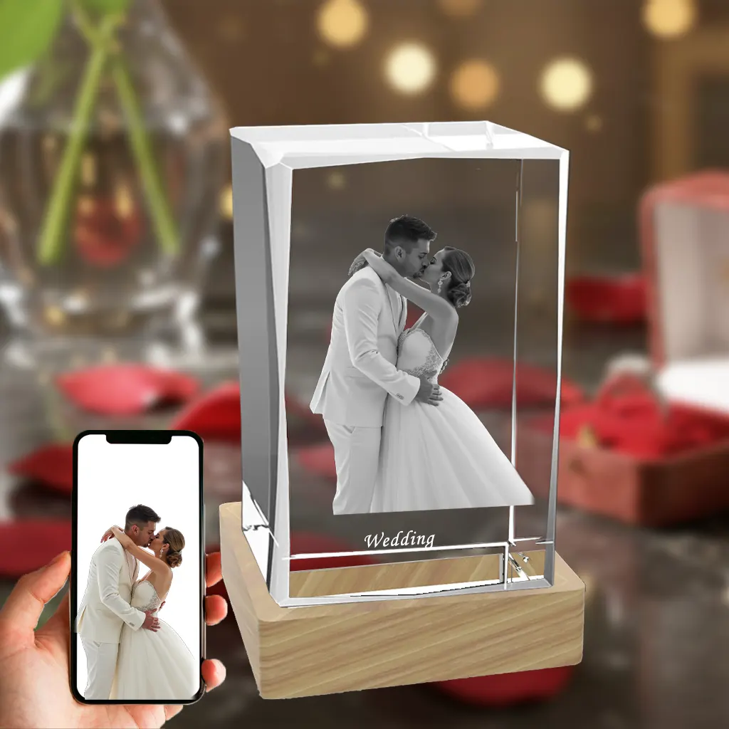 A vertical beveled rectangular 3D crystal, internally engraved with a romantic wedding photo of a couple kissing, with the text "Wedding" below. The crystal sits on an illuminated wooden LED base against a romantic red rose background, with a hand in the bottom-left holding a phone showing the original color photo.