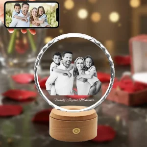A 3D photo of a happy family of four is engraved inside a sunflower-shaped crystal with diamond-cut edges. The crystal sits on an illuminated wooden base, surrounded by red rose petals, making it a perfect Valentine's Day gift. A phone in the corner shows the original color photograph.