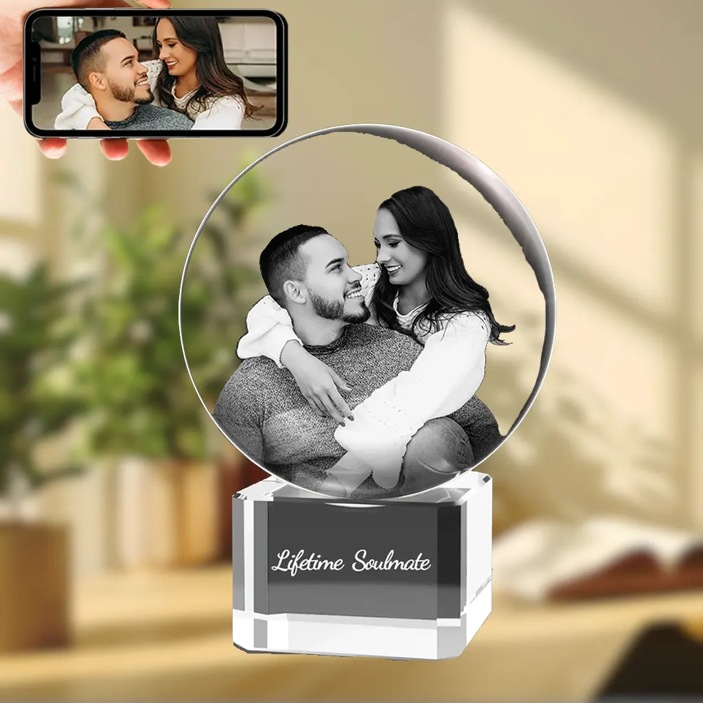 A YiYues personalized 3D crystal ball featuring a custom photo of a romantic couple, on a square crystal base engraved with 'Lifetime Soulmate'. A hand holds a smartphone showing the original color source photo.