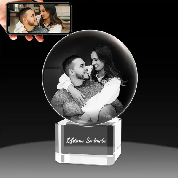 A YiYues personalized 3D crystal ball featuring a lifelike 3D portrait of a couple engraved inside. It sits on a crystal base inscribed with 'Lifetime Soulmate'. A smartphone shows the original 2D photo.