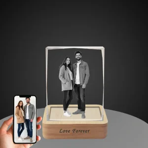 YiYues personalized 3D deckle-edge crystal block featuring a full-body custom photo of a couple, with 'Love Forever' engraved on the lighted wooden base, shown next to a smartphone displaying the original color photo.