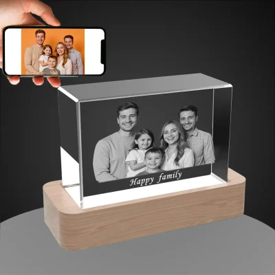 A personalized horizontal YiYues 3D Rectangular Crystal on a lighted wooden base, featuring a custom family photo and the text 'Happy family'. A hand holds a smartphone showing the original color photo.