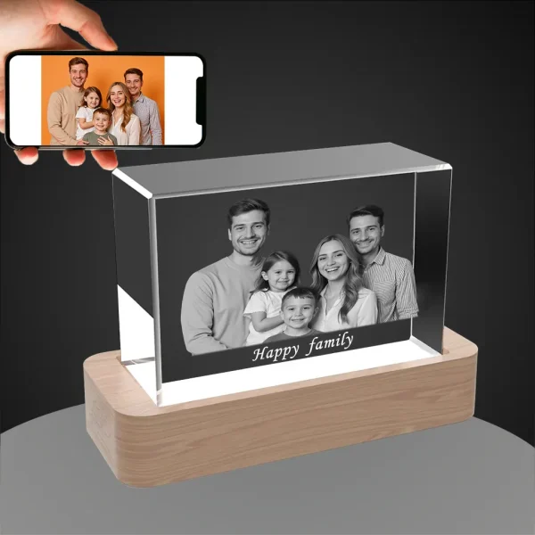 A personalized horizontal YiYues 3D Rectangular Crystal on a lighted wooden base, featuring a custom family photo and the text 'Happy family'. A hand holds a smartphone showing the original color photo.