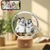 A personalized YiYues 3D Sunflower Crystal showcasing a custom family portrait, with 'Family Joyous Moments' engraved. It rests on a lighted wooden base, with a smartphone displaying the original color photo used for the engraving.