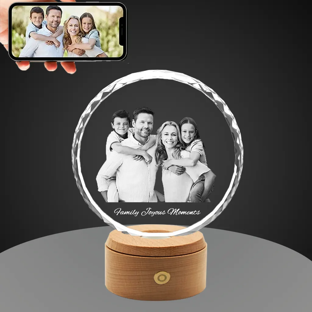 A YiYues personalized 3D Sunflower Crystal featuring a custom photo of a happy family, engraved with 'Family Joyous Moments', on a lighted wooden base. A smartphone shows the original color source photo.